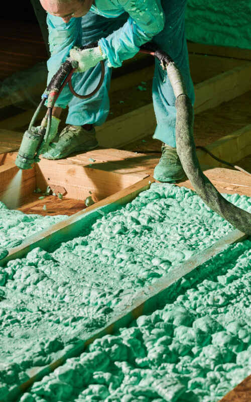 Male builder insulating wooden frame house. Cropped view of man worker spraying polyurethane foam on floor inside of future cottage, using plural component gun. Construction and insulation concept.