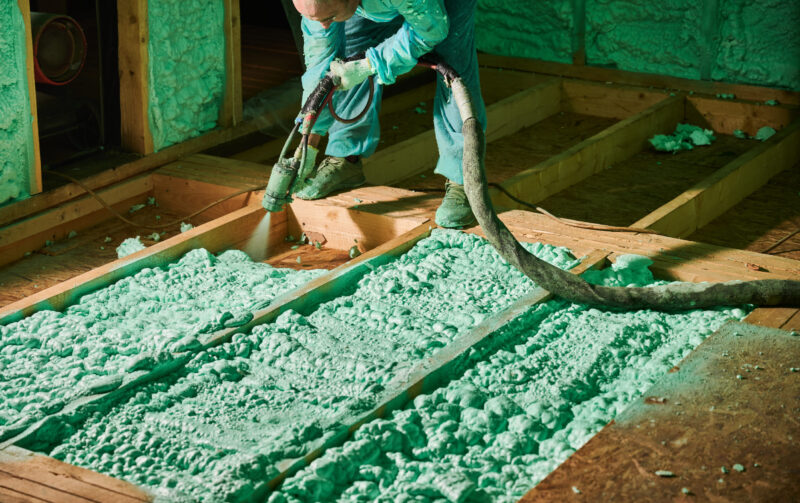 Male builder insulating wooden frame house. Cropped view of man worker spraying polyurethane foam on floor inside of future cottage, using plural component gun. Construction and insulation concept.