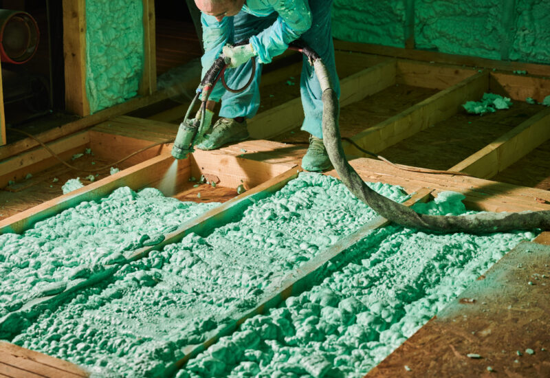 Male builder insulating wooden frame house. Cropped view of man worker spraying polyurethane foam on floor inside of future cottage, using plural component gun. Construction and insulation concept.